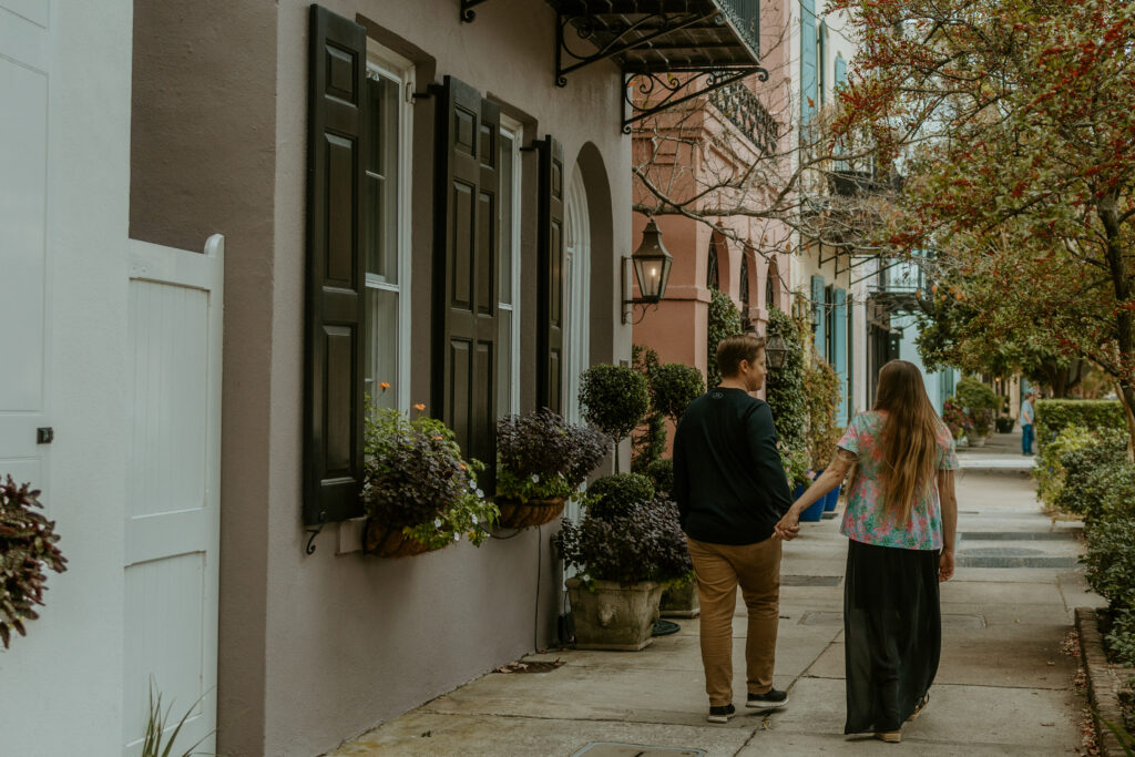  couple strolling hand in hand down Rainbow Row in downtown Charleston, surrounded by vibrant pastel-colored historic homes and lush greenery, capturing a romantic and timeless moment."