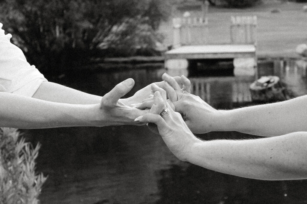 A candid documentary-style photo of a couple standing in front of a peaceful pond, their reflections shimmering in the water. Surrounded by lush greenery and soft natural light, the image captures their authentic connection and the serene beauty of the moment.