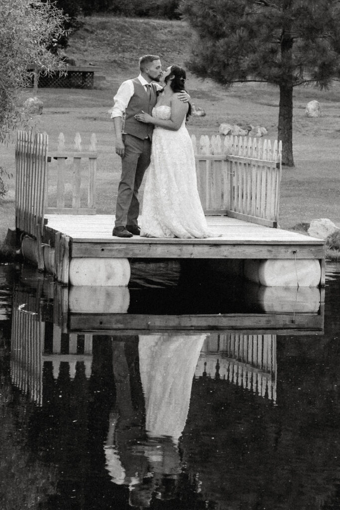 A documentary-style photo of a couple standing near a tranquil pond, with their reflection perfectly mirrored in the water. The scene captures the couple’s intimate connection, framed by the serene natural surroundings and soft, ambient light, creating a timeless and artistic moment.